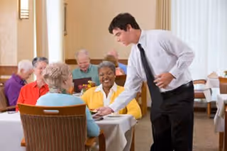 A server in a white shirt and black tie serves smiling elderly residents seated at a table in a bright dining room.