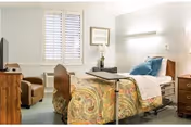 A furnished senior bedroom with an adjustable hospital-style bed covered in a paisley bedspread, a tray table, armchair, TV, and a window with shutters.