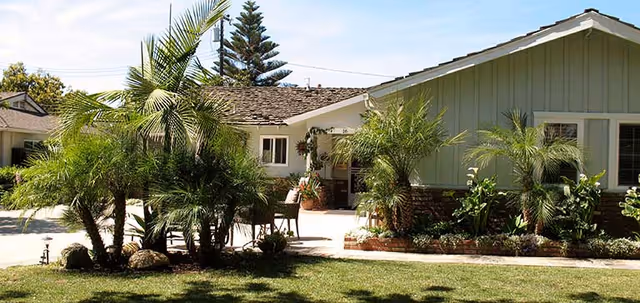 Exterior view of a single-story residential building with a well-maintained garden featuring various palm trees and shrubs. The building has light-colored siding and a shingled roof, with a small patio area visible near the entrance.