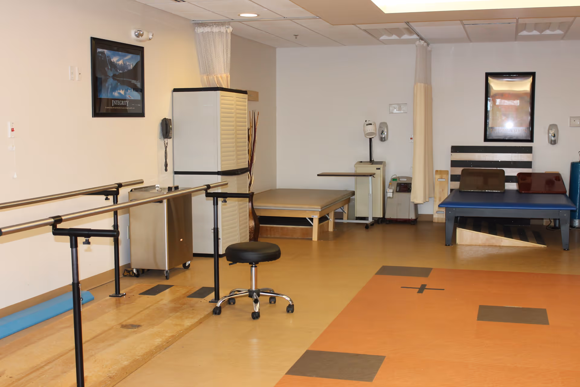 Physical therapy room with parallel bars for walking exercises, a black rolling stool, therapy tables, and medical equipment against beige walls and a patterned floor.