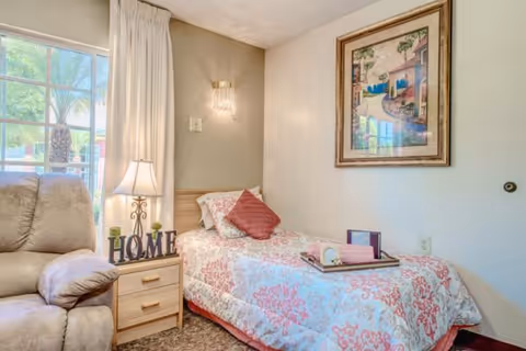 Cozy single bedroom with a twin bed dressed in patterned bedding and pillows, a nightstand with a lamp and 'HOME' letters, and a recliner by a window.