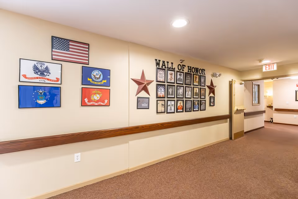 A hallway in an assisted living facility with a 'Wall of Honor' display featuring framed photos and plaques honoring military veterans. The wall also has several military flags and two decorative stars. The hallway has beige walls, brown carpet, and a wooden handrail along the wall. An exit sign is visible above an open door at the end of the hallway.