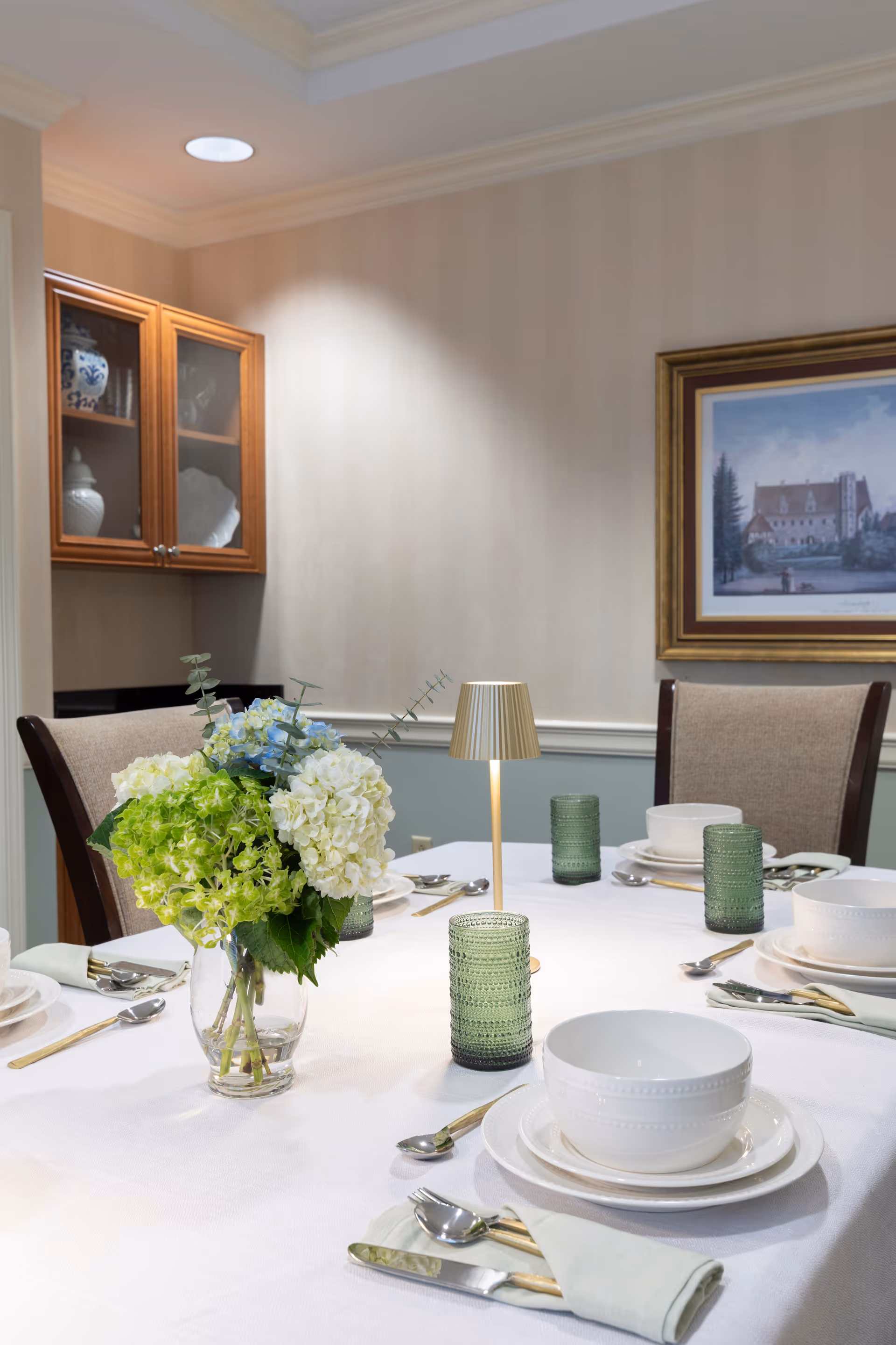 A neatly set dining table with white tablecloth, white bowls and plates, silver cutlery, green textured drinking glasses, and a small gold table lamp. A vase with green and white hydrangea flowers is placed in the center. The background shows beige wallpaper, a framed painting, and a wooden cabinet with glass doors containing decorative items.