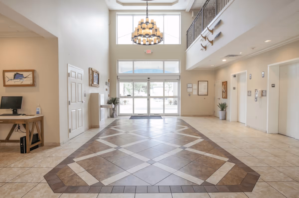 Bright and spacious senior living facility lobby with high ceilings, large chandelier, tiled floor with decorative pattern, glass entrance doors, a computer desk on the left, and elevator doors on the right.