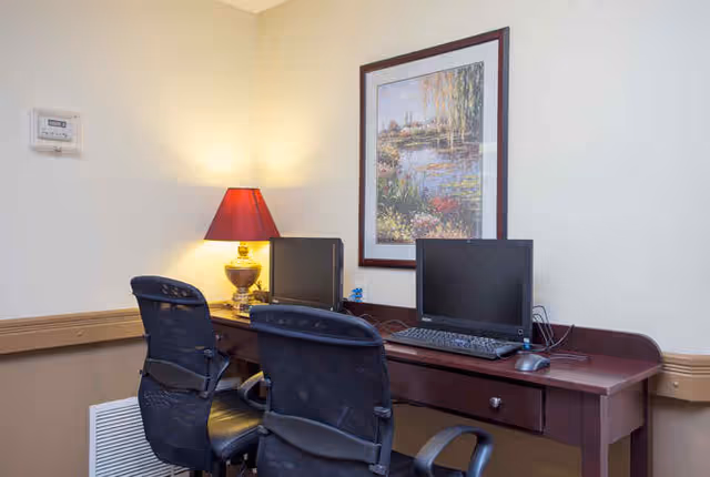 A wooden desk with two computer monitors and office chairs beneath a framed landscape painting and a lit table lamp.