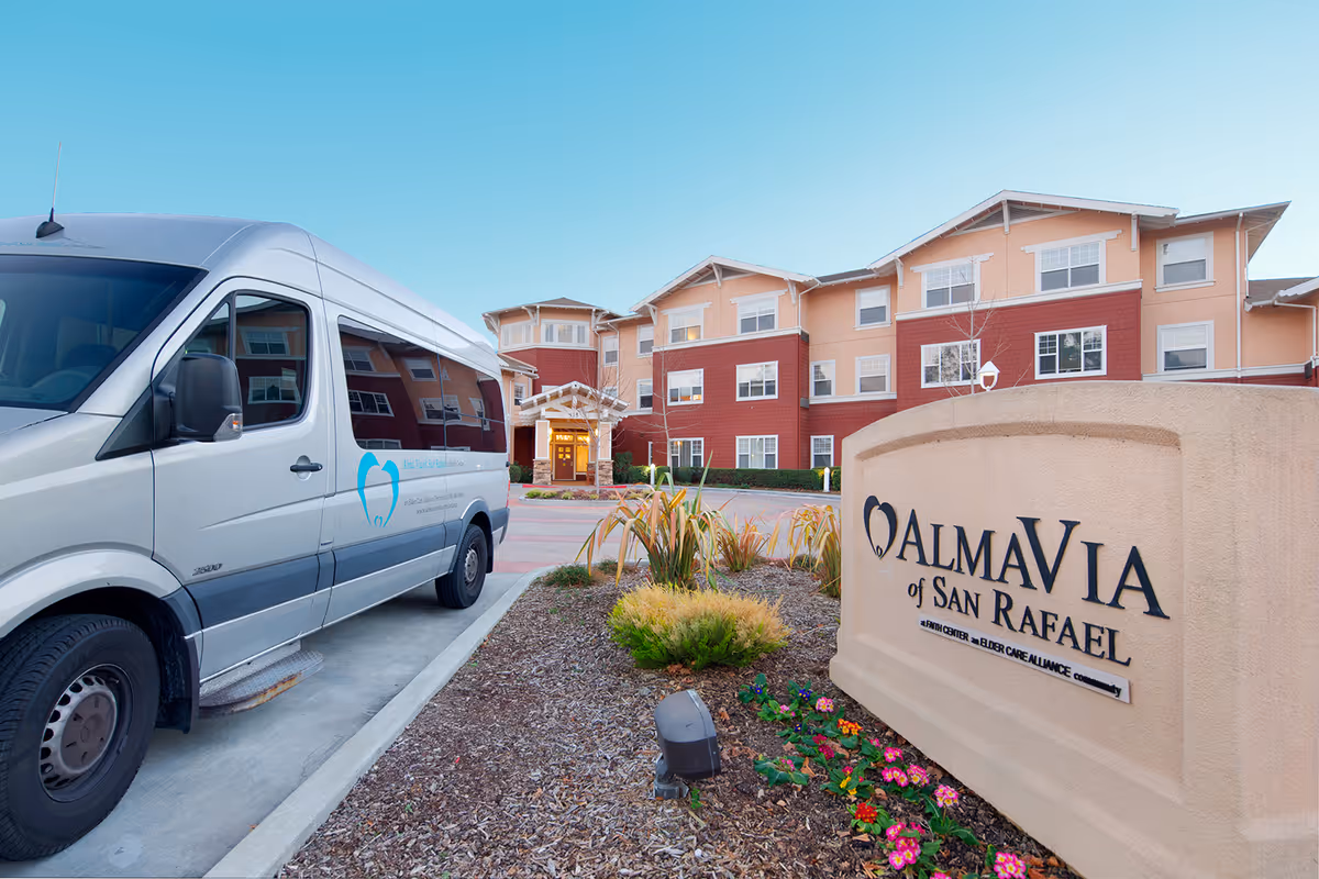 Exterior view of AlmaVia of San Rafael senior living facility with a beige and red multi-story building in the background, a white shuttle van parked on the left, and a large sign with the facility name in the foreground surrounded by landscaping and flowers.