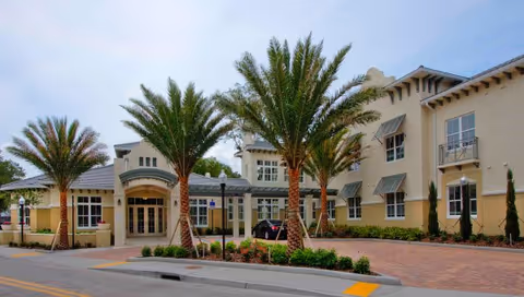 Exterior view of a senior living facility named The Estate at Hyde Park, featuring a beige building with multiple windows, palm trees lining the entrance, and a paved driveway under a partly cloudy sky.