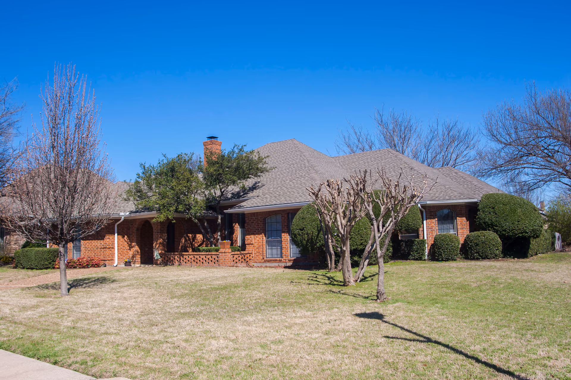 A single-story brick house with a gray shingled roof, surrounded by trimmed bushes and leafless trees on a grassy lawn under a clear blue sky.