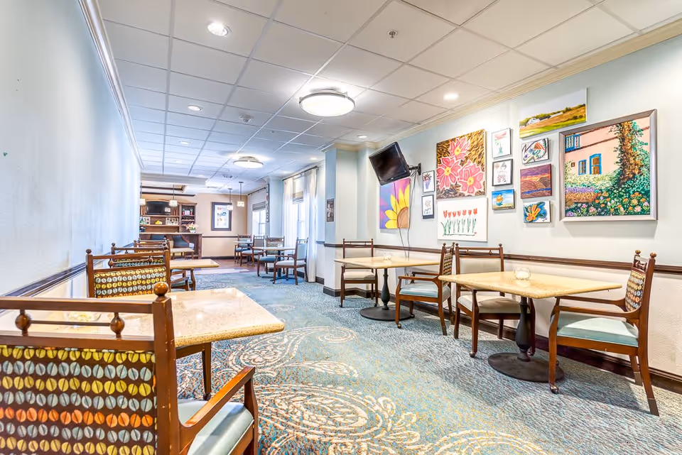 A bright dining room in Sierra Blanca Active Adult Living featuring multiple square tables with wooden chairs that have patterned cushions. The room has a patterned carpet, light blue walls, and several colorful paintings on one wall. A flat-screen TV is mounted on the wall above the paintings. Large windows with curtains allow natural light to enter the space.