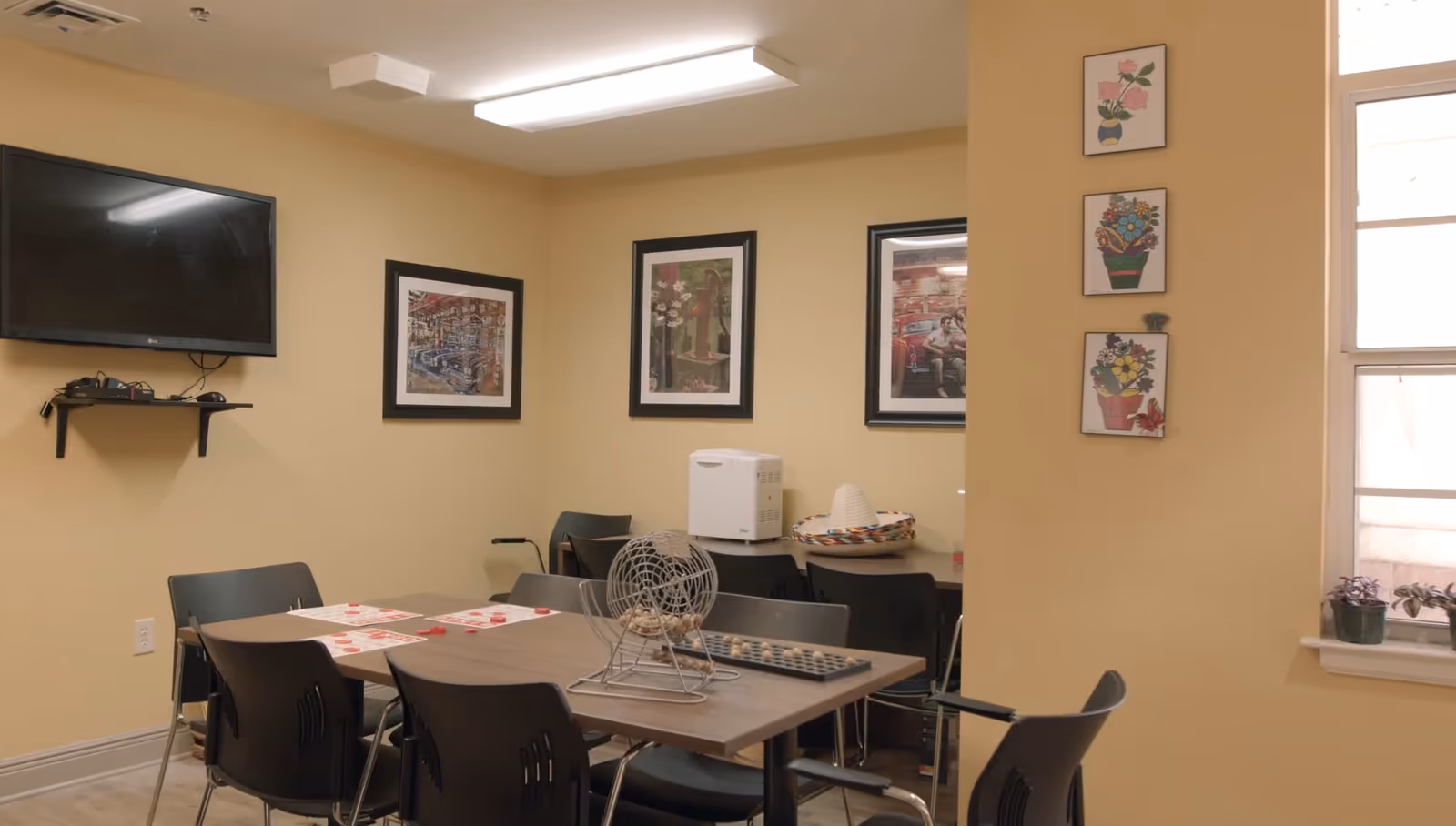 A small activity room with a table and several black chairs around it. On the table, there is a bingo cage with balls and bingo cards. The walls are painted beige and decorated with framed pictures and floral artwork. A flat-screen TV is mounted on the wall above a small shelf holding electronic devices. A window with potted plants on the sill is visible on the right side.