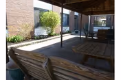 Covered outdoor seating area with wooden benches and tables, adjacent to a brick building with windows and some greenery along the building's edge.