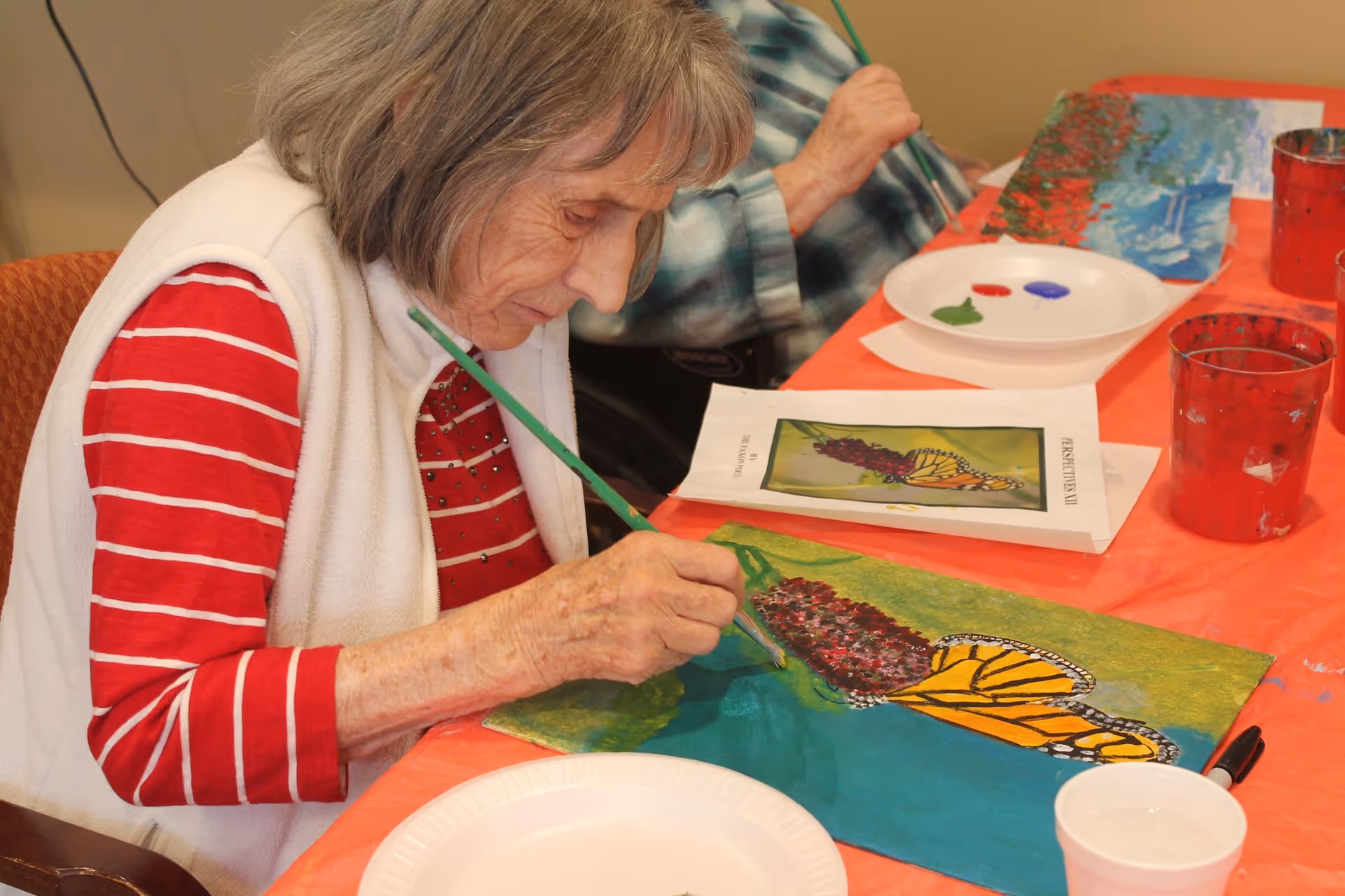 An elderly woman wearing a red and white striped shirt and white vest is painting a colorful picture of a butterfly on a flower at a table covered with an orange tablecloth. There are paint supplies, including a palette with red, blue, and green paint, and cups on the table. Another person is partially visible in the background also engaged in painting.