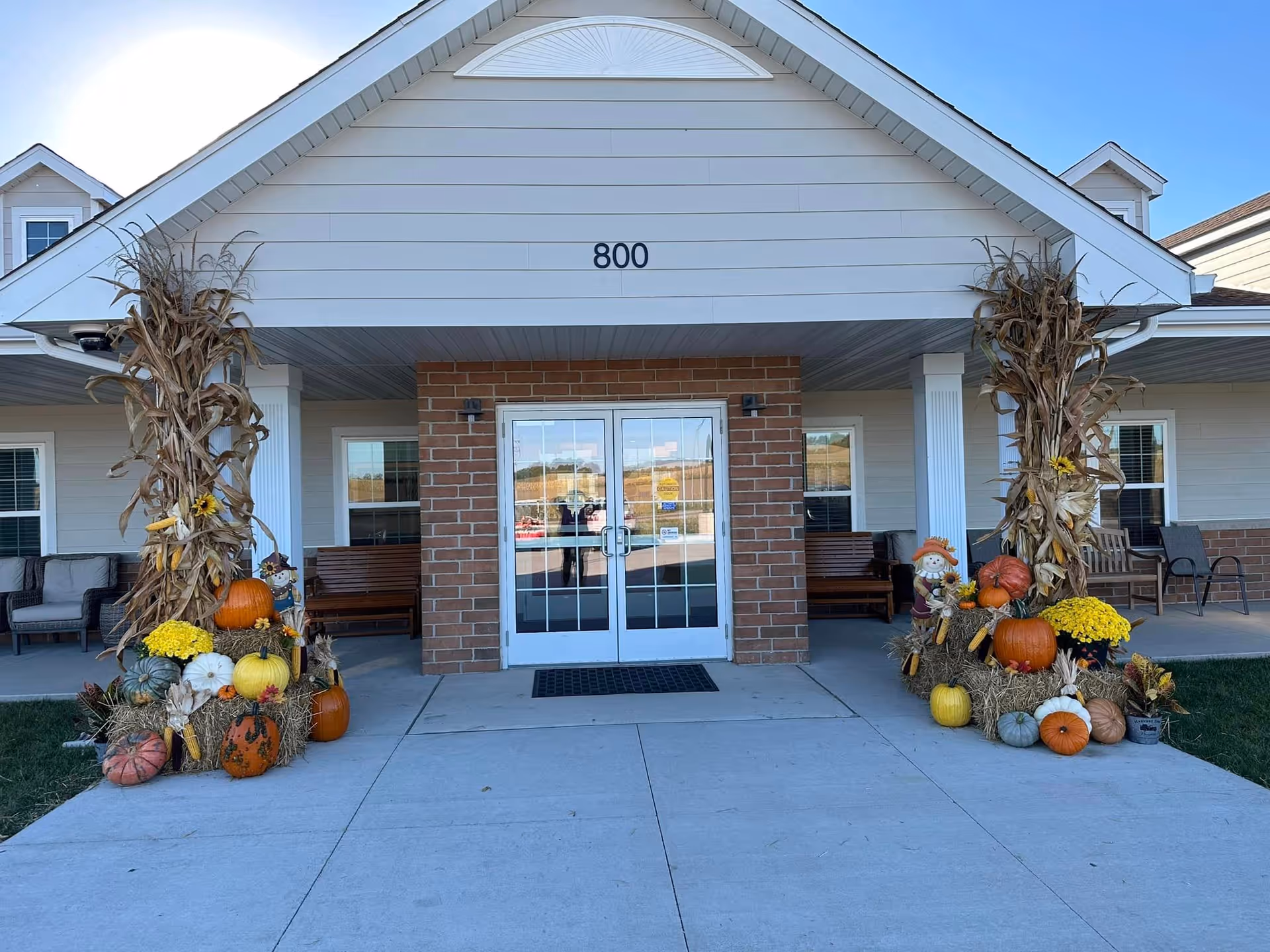 Entrance of a senior living community building with the number 800 above double glass doors. The entrance is decorated with autumn-themed arrangements including pumpkins, hay bales, corn stalks, yellow flowers, and small scarecrow figures on either side of the doorway. There are benches and chairs on the porch area under the roof.