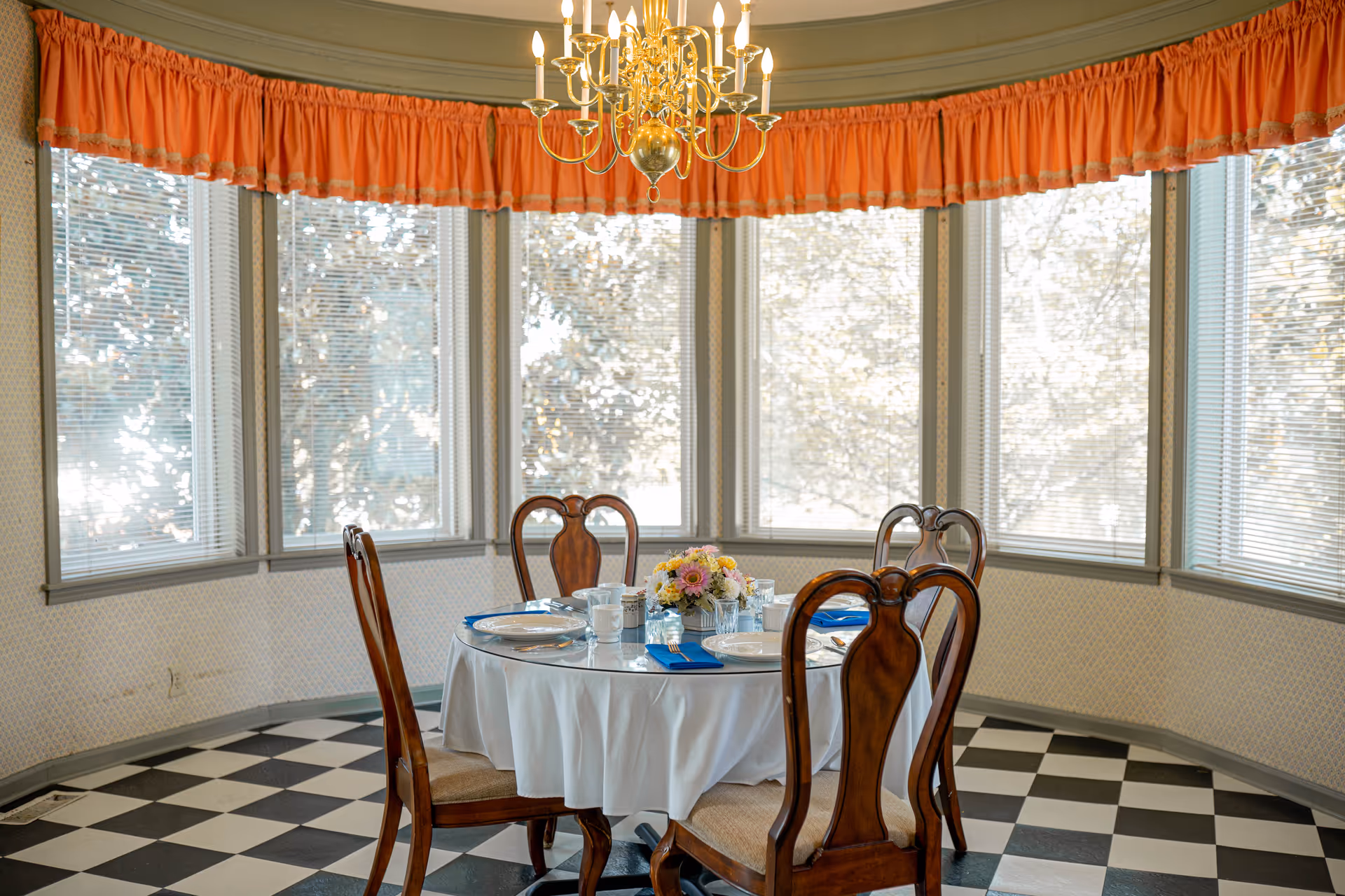 A dining area with a round table covered with a white tablecloth, set with plates, glasses, mugs, and blue napkins. Four wooden chairs surround the table. A floral centerpiece is on the table. The room has large windows with blinds and orange valance curtains, a black and white checkered floor, and a brass chandelier hanging above the table.