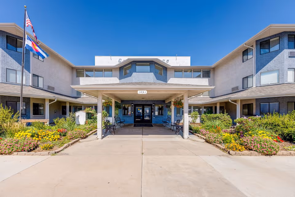 Front exterior view of Greeley Place | Sky Active Living facility with a covered entrance, benches, flower beds on both sides, and two flagpoles displaying the American and Colorado state flags under a clear blue sky.