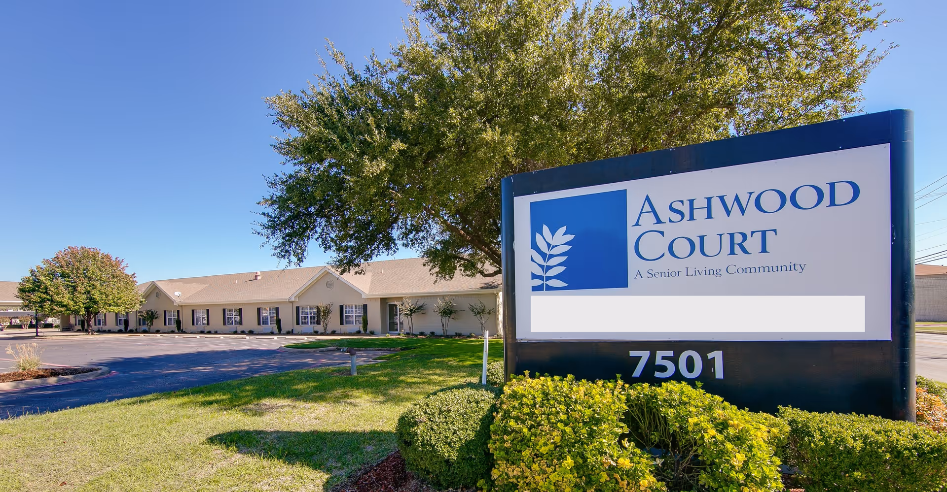 Exterior view of Ashwood Court senior living community building with a large sign displaying the facility name and address number 7501, surrounded by green bushes and trees under a clear blue sky.