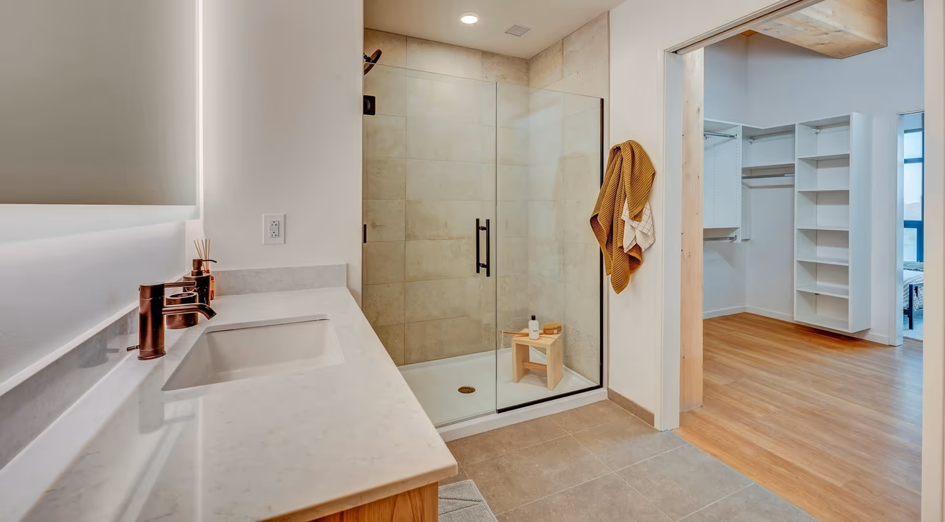 Modern bathroom with a glass-enclosed shower featuring beige tiles, a small wooden stool with toiletries inside the shower, a white countertop with a built-in sink and bronze faucet, and a doorway leading to a walk-in closet with wooden flooring and white shelving.