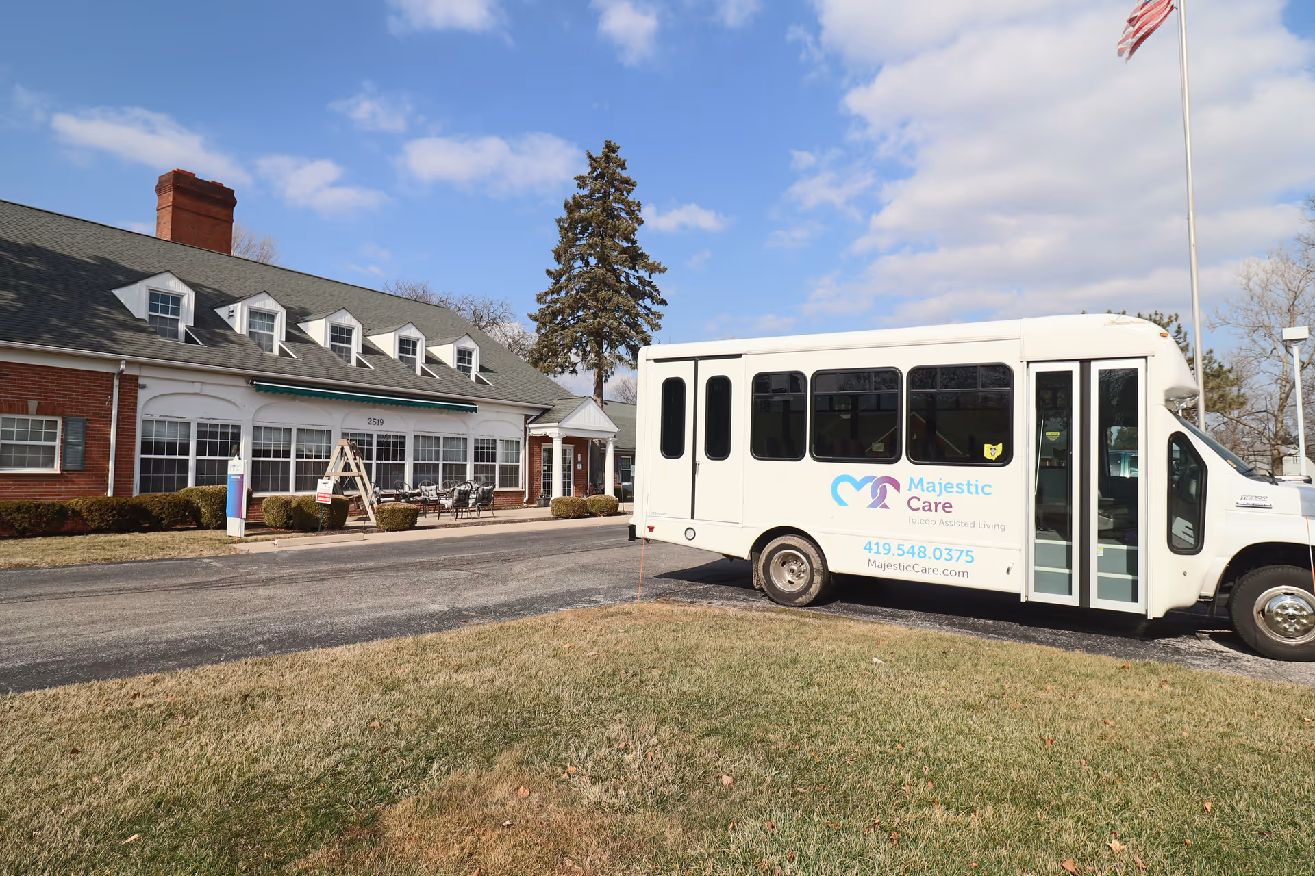 Exterior view of Majestic Care of Toledo Assisted Living building with a white shuttle bus parked in front. The building has a brick facade, multiple windows, and a green awning. There is a flagpole with an American flag on the right side of the image and a clear blue sky with some clouds.