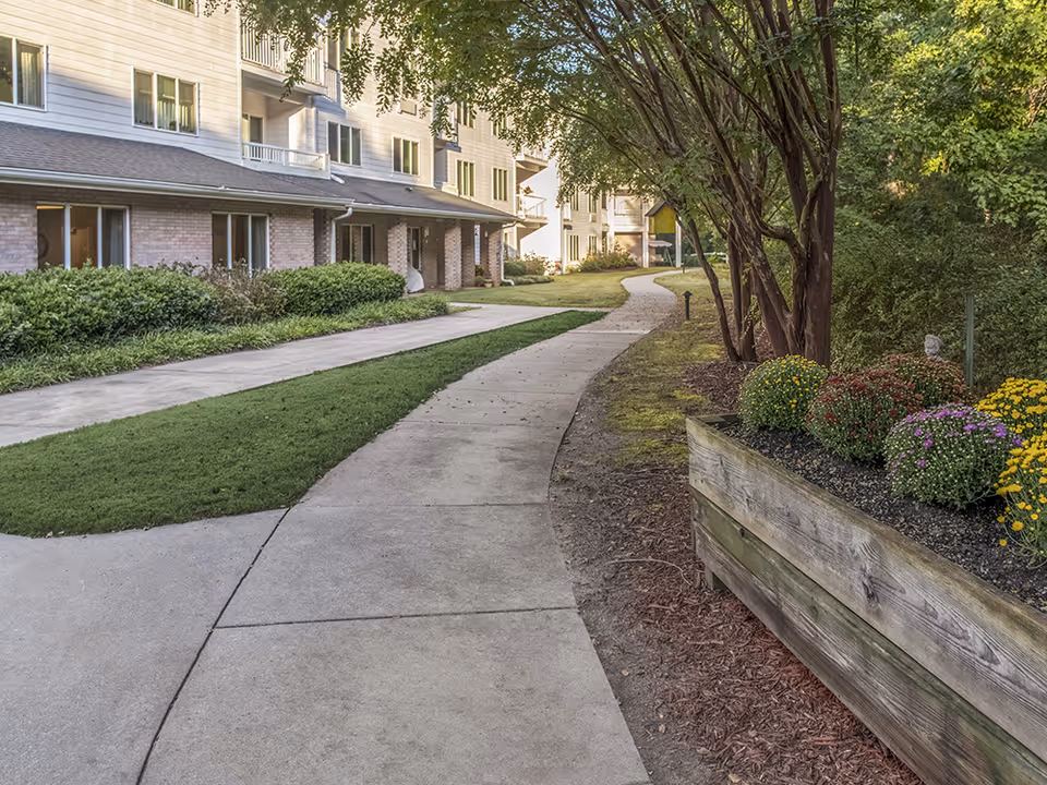 A winding concrete pathway through a landscaped garden area with green grass, bushes, and colorful flowers in a wooden planter. The path runs alongside a multi-story residential building with windows and balconies, surrounded by trees and greenery.