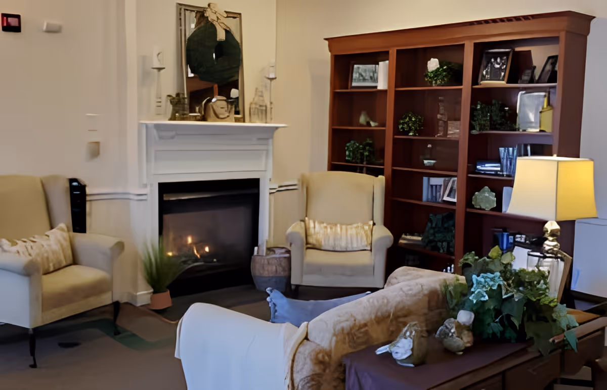 Cozy living room with a lit fireplace, two beige armchairs with decorative pillows, a large wooden bookshelf filled with plants and framed photos, a beige couch with a white throw, and a table lamp on a side table.