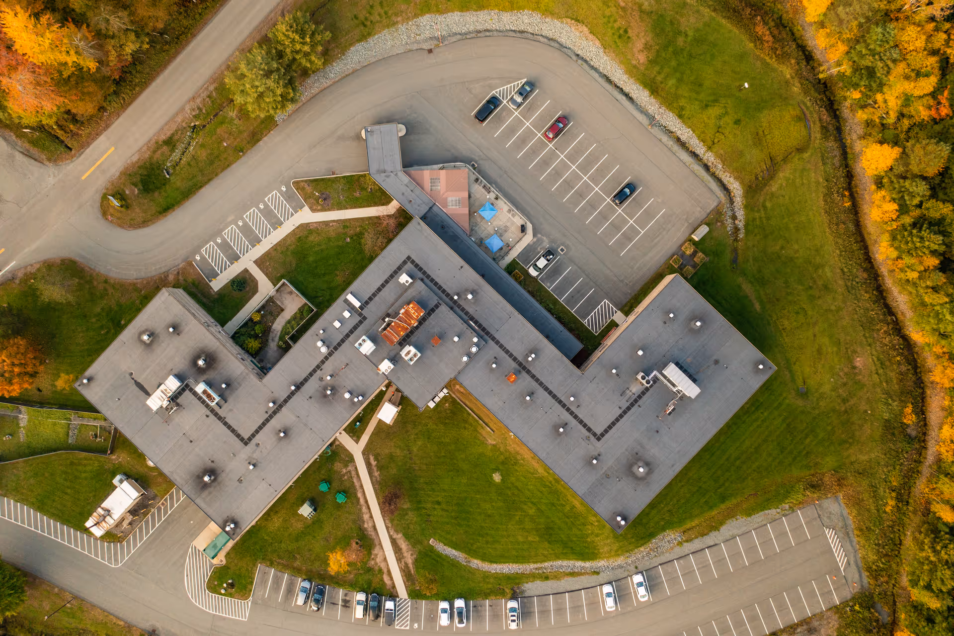 Aerial view of the Care Center at Sunset Lake showing the building's roof, surrounding parking lots with cars, green lawns, and nearby trees with autumn foliage.
