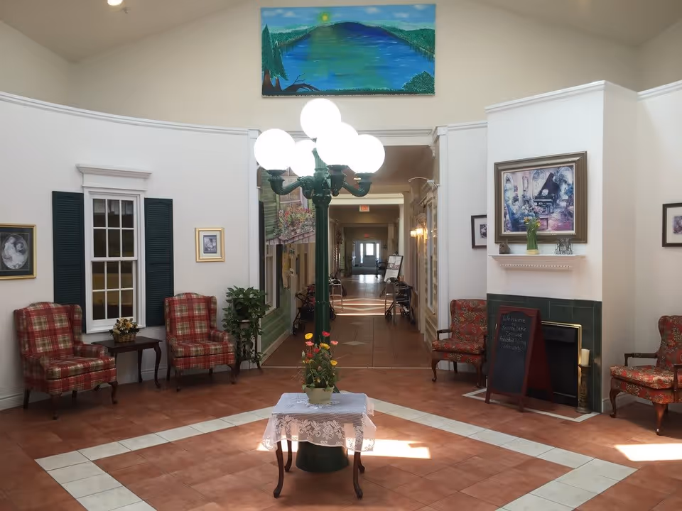 A bright and spacious common area in a senior living facility with a central vintage-style lamp post featuring multiple glowing globes. The room has tiled floors with a white border, several upholstered armchairs with red and plaid patterns, a small table with a lace cloth and a flower pot, and framed artwork on the walls. A hallway extends from the room, with walkers visible along the sides.
