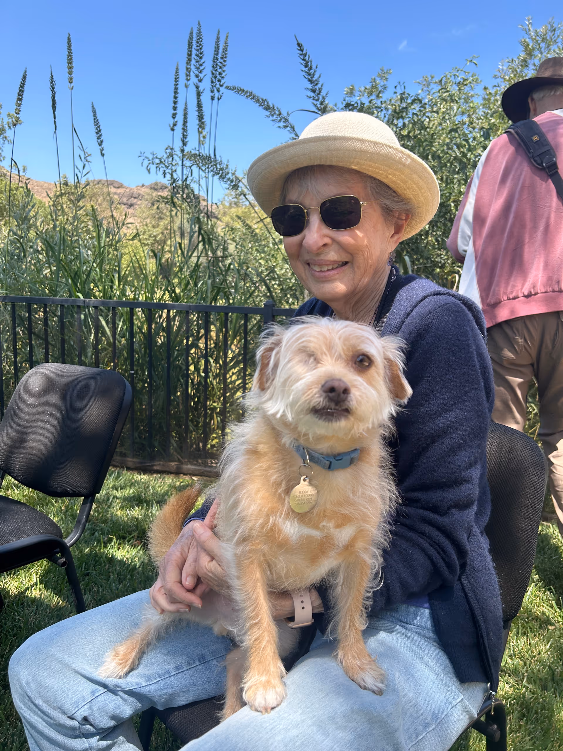 An elderly woman wearing a beige hat, sunglasses, a navy blue sweater, and light blue jeans is sitting outdoors on a black chair, holding a small light brown dog with a blue collar. Behind them is greenery, including tall plants and trees, and a clear blue sky.