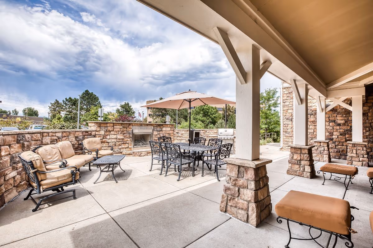 Covered outdoor patio with stone pillars, cushioned lounge chairs, a metal dining set with umbrella, and a stone privacy wall with a built-in fireplace.