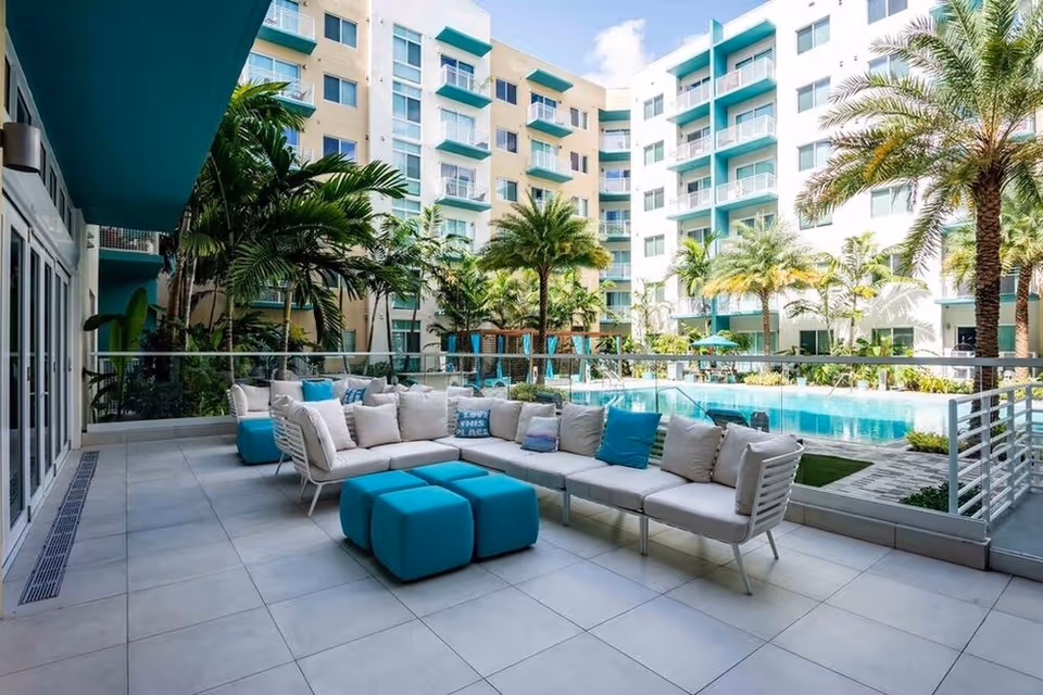 Outdoor patio area with modern white and turquoise cushioned seating and ottomans, overlooking a swimming pool surrounded by palm trees and a multi-story residential building in the background under a partly cloudy sky.