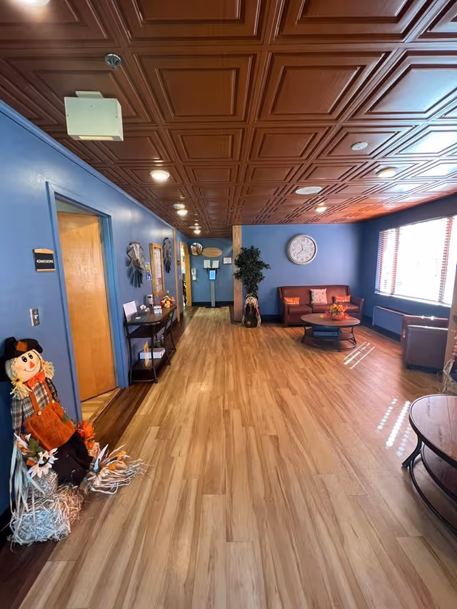 Long reception-style common area with wood-pattern flooring, blue walls, paneled ceiling, seating area, and autumn decorations.