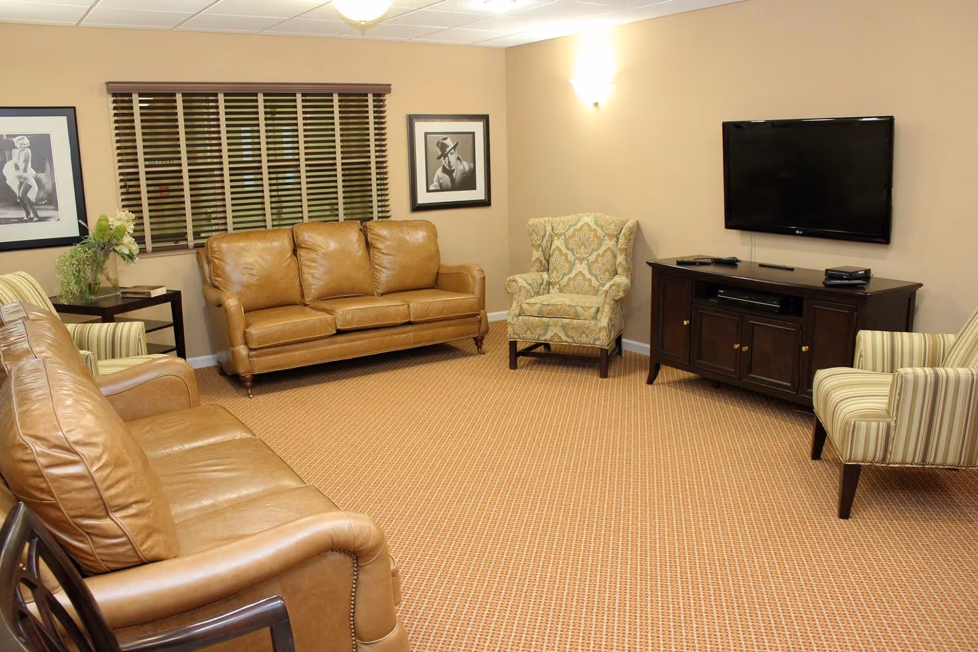 A cozy living room with two brown leather sofas, two patterned armchairs, a wooden TV stand with a flat-screen TV mounted on the wall above it, and framed black and white photographs on the walls. The room has beige walls, a window with wooden blinds, and a carpeted floor.