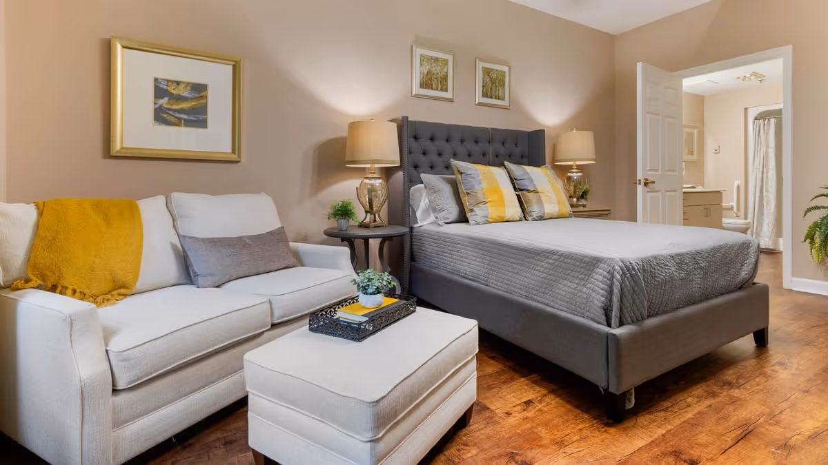 A cozy bedroom featuring a gray upholstered bed with decorative pillows, two bedside tables with lamps, a white sofa with a yellow throw blanket and gray pillow, and a matching ottoman with a tray. The room has wooden flooring and beige walls with framed artwork. An open door reveals a bathroom in the background.