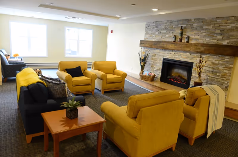 A cozy living room area with four mustard yellow armchairs and a dark blue sofa arranged around a wooden coffee table with a small plant. The room features a stone fireplace with a wooden mantel decorated with vases and a basket with magazines. Large windows allow natural light to fill the space.