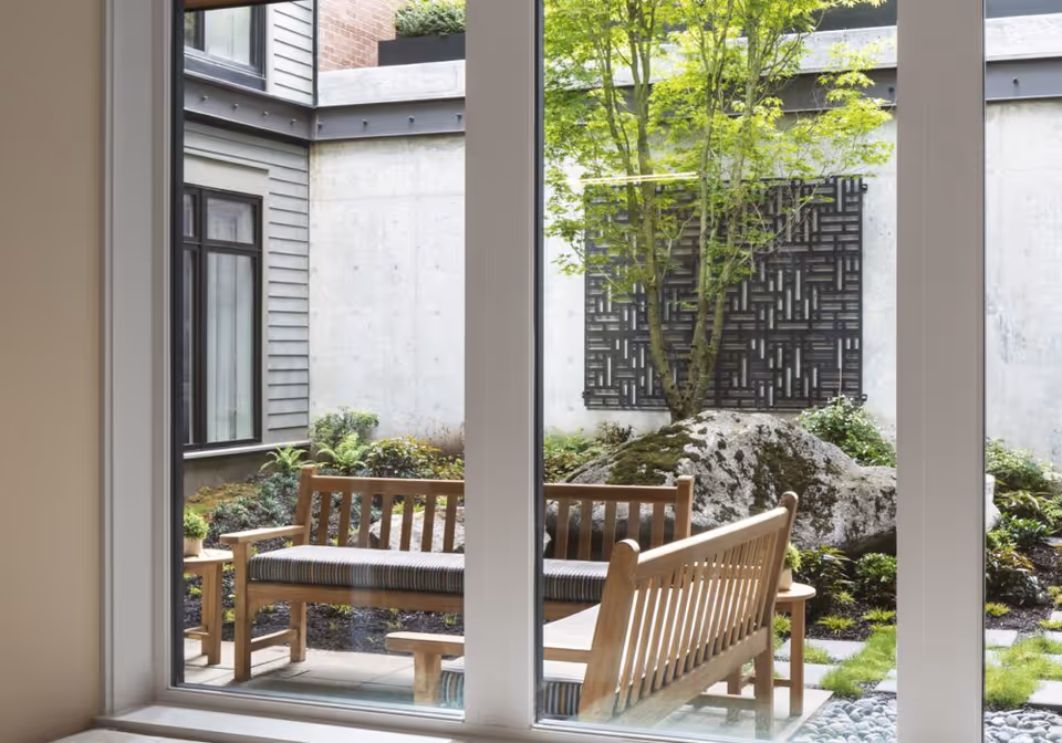 View through a large window showing an outdoor courtyard with wooden benches, a small table, a large rock, a tree, and decorative wall art on a concrete wall surrounded by plants.