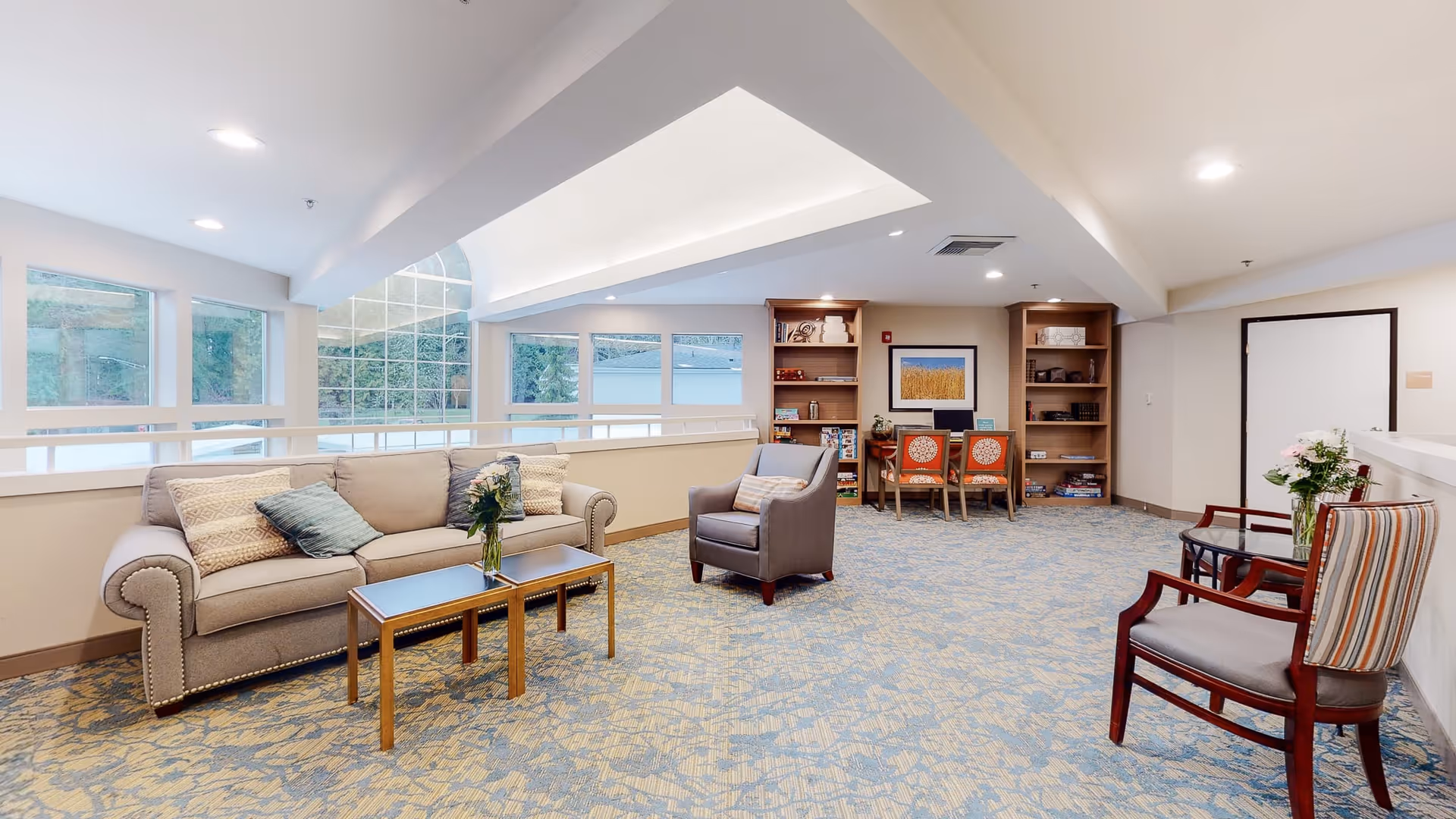 A bright and spacious common area with a beige sofa, an armchair, and two wooden chairs with striped cushions. There is a coffee table with a vase of flowers in front of the sofa. The room has large windows letting in natural light, built-in bookshelves with books and decorative items, and a carpeted floor with a blue and beige pattern.