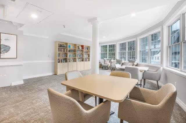 A bright and spacious common area in a senior living facility featuring a square wooden table with four upholstered chairs in the foreground. In the background, there is a large bookshelf filled with books and decorative items, and a row of comfortable armchairs positioned along a wall of large windows letting in natural light.