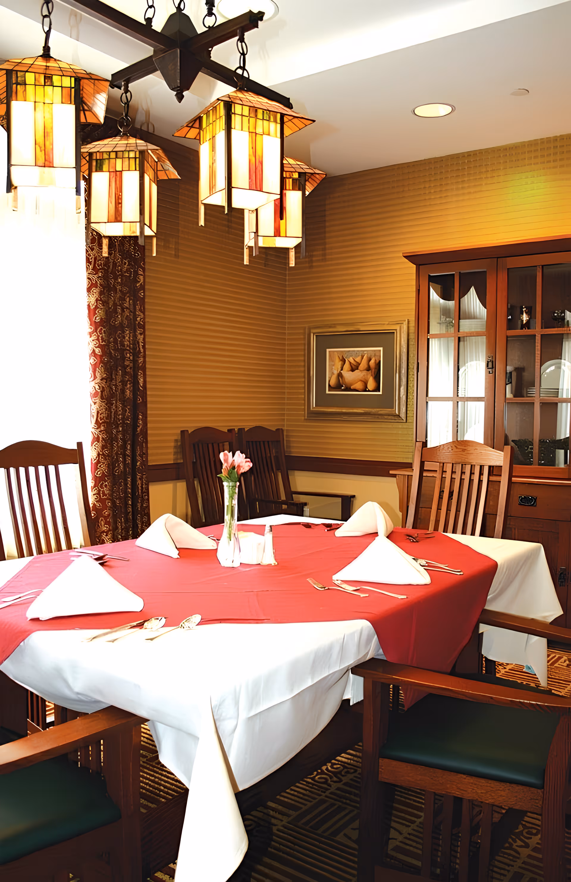 A dining room with a wooden table covered by a white tablecloth and a red table runner, set with folded white napkins and silverware. The room features wooden chairs, a wooden cabinet with glass doors, a framed picture on the wall, patterned curtains, and hanging stained glass pendant lights.