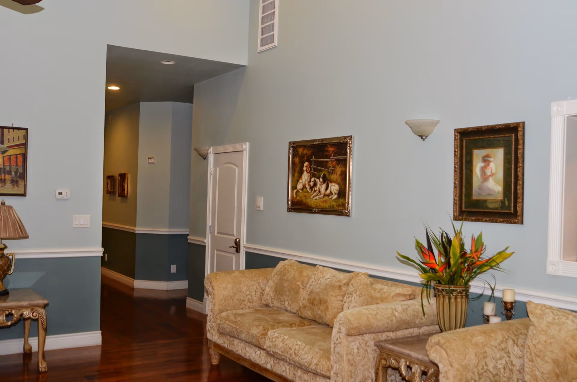 A sitting area with upholstered sofas, framed paintings on a pale blue wall, a side table with a lamp and a vase of flowers, and a hallway leading deeper into the facility.