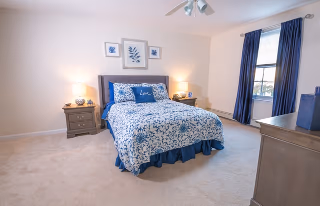 Bright bedroom with a queen bed in blue floral bedding, two nightstands with lamps, a dresser, and a window with dark blue curtains.