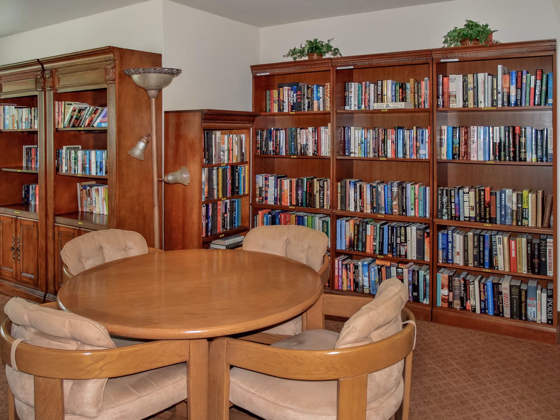 A cozy reading room with wooden bookshelves filled with books along the walls. In the center, there is a round wooden table surrounded by four cushioned chairs. A floor lamp with multiple light fixtures stands beside the bookshelves.