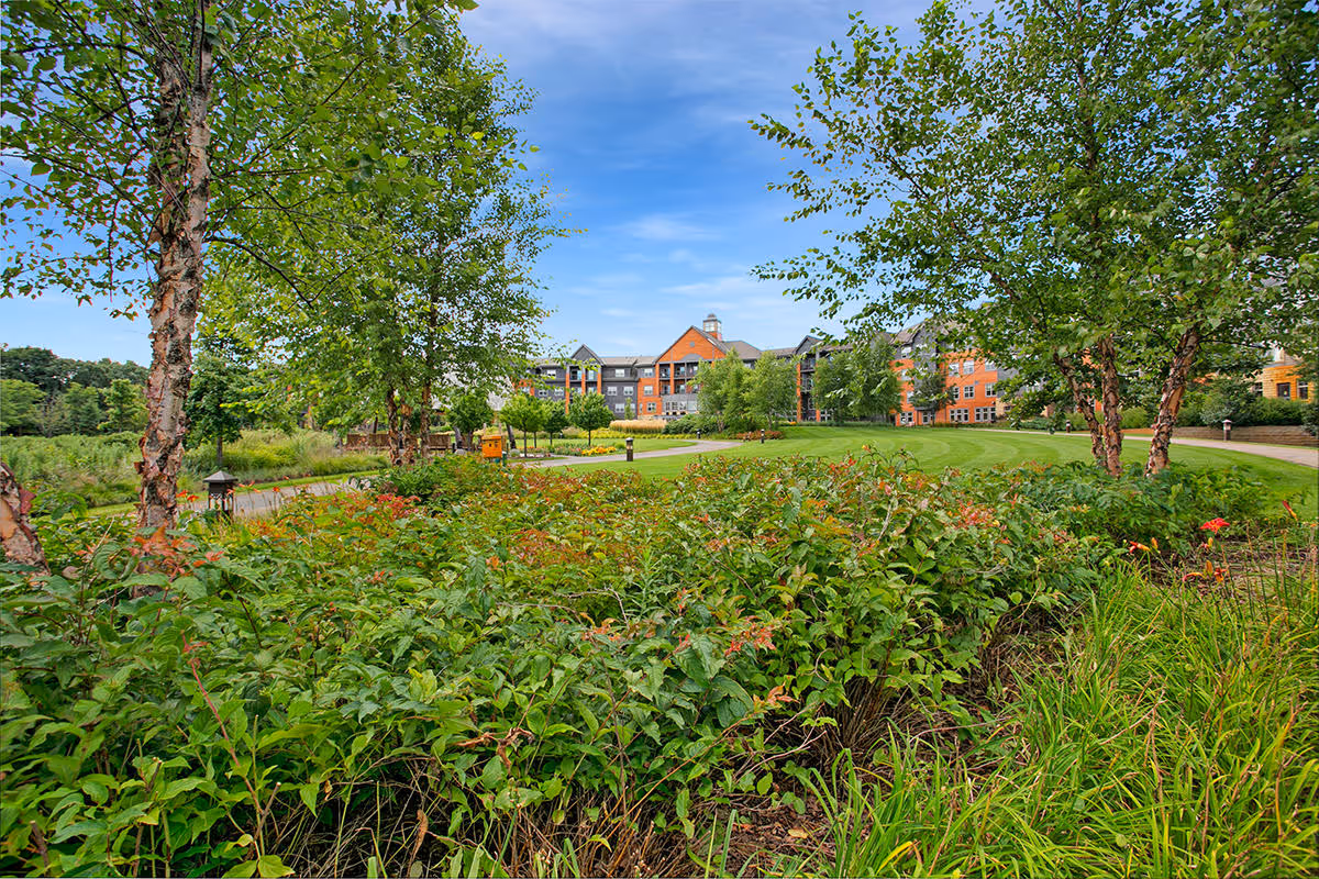 Landscaped garden and lawn with trees and shrubs in front of a multi-story brick residential building under a blue sky.