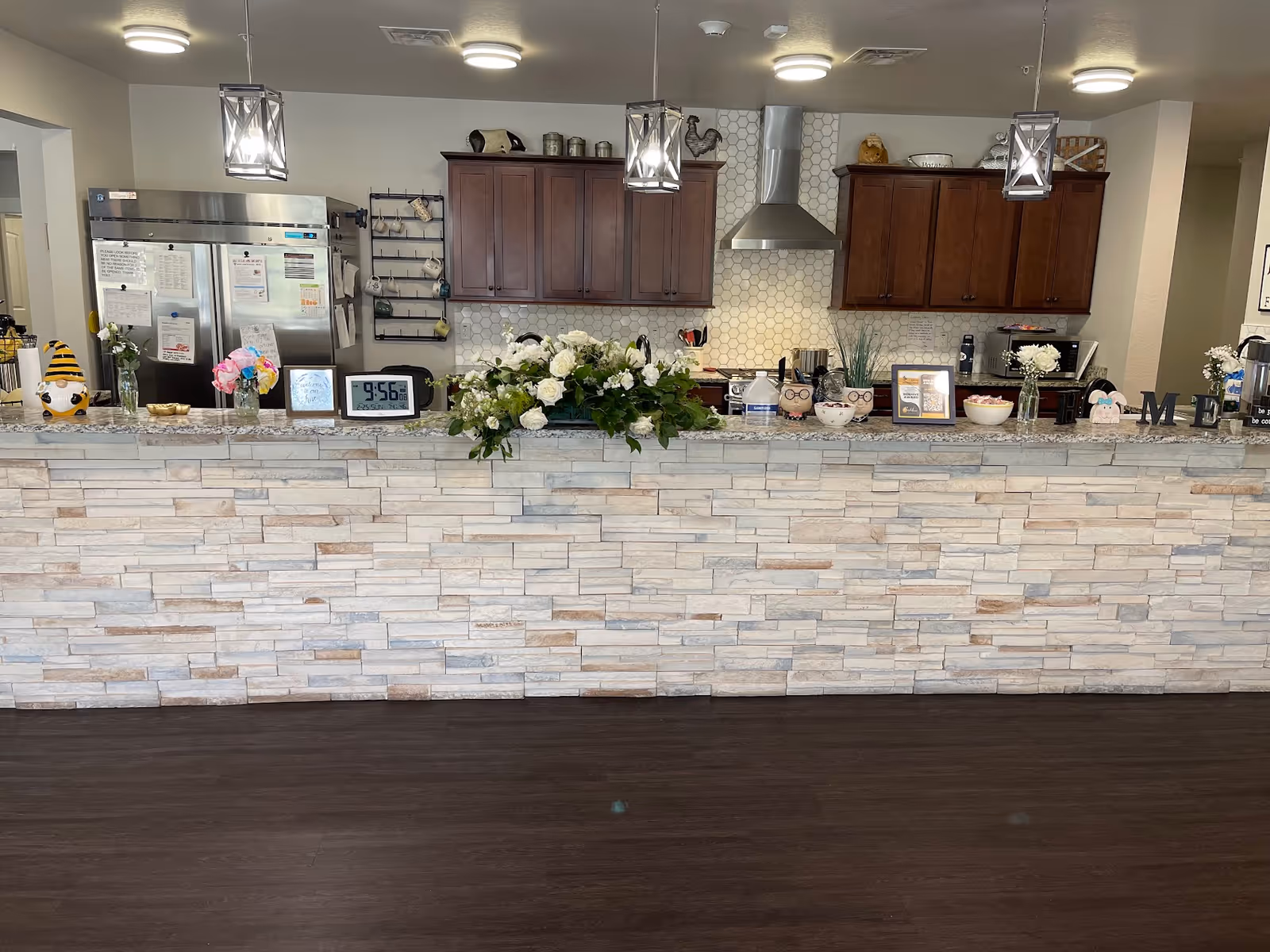 Interior view of a kitchen area in an assisted living facility with a stone-faced counter in the foreground. The counter is decorated with flowers, small decorative items, and a digital clock showing 9:56. Behind the counter are wooden cabinets, a stainless steel refrigerator, a stove with a hood, and various kitchen utensils. Three pendant lights hang from the ceiling above the counter.