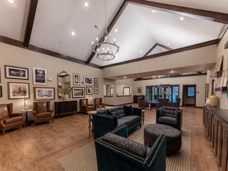 A spacious senior living facility common area with high vaulted ceilings and exposed wooden beams. The room features a chandelier, several framed pictures on the walls, and a mix of seating including dark green upholstered sofas and chairs, brown leather armchairs, and a round ottoman. There is a wooden floor and a large rug under the central seating area. The back of the room has a reception desk and glass doors leading outside.