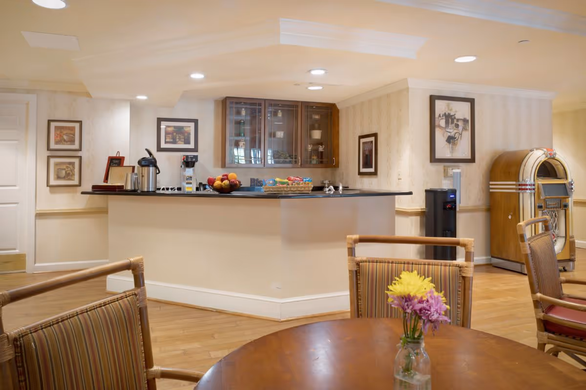 A cozy common area with a wooden round table and chairs in the foreground, a counter with snacks and a coffee station in the background, framed pictures on the walls, a water dispenser, and a vintage jukebox in the corner.