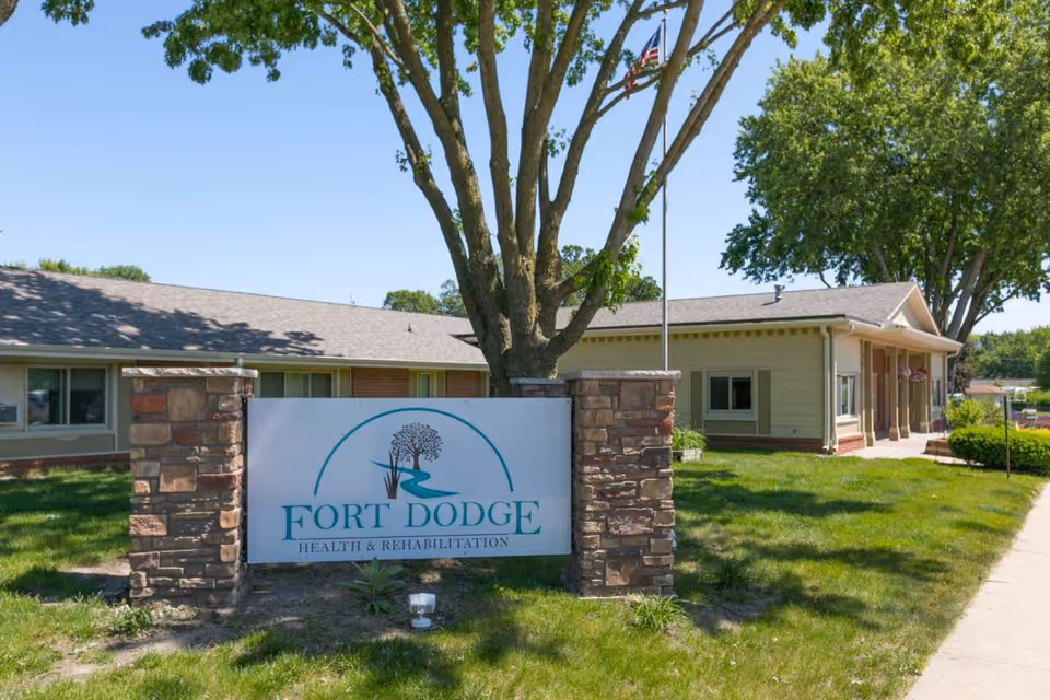 Exterior view of Fort Dodge Health and Rehabilitation facility with a large sign in front supported by two stone pillars, a tree behind the sign, and an American flag on a flagpole. The building is single-story with beige siding and a shingled roof, surrounded by green grass and trees under a clear blue sky.