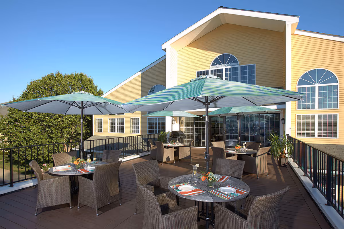 Outdoor patio area at The Village at Willow Crossings with several round tables and wicker chairs under large green and white striped umbrellas. The patio is elevated with black railings and is adjacent to a yellow building with large windows. The sky is clear and blue.