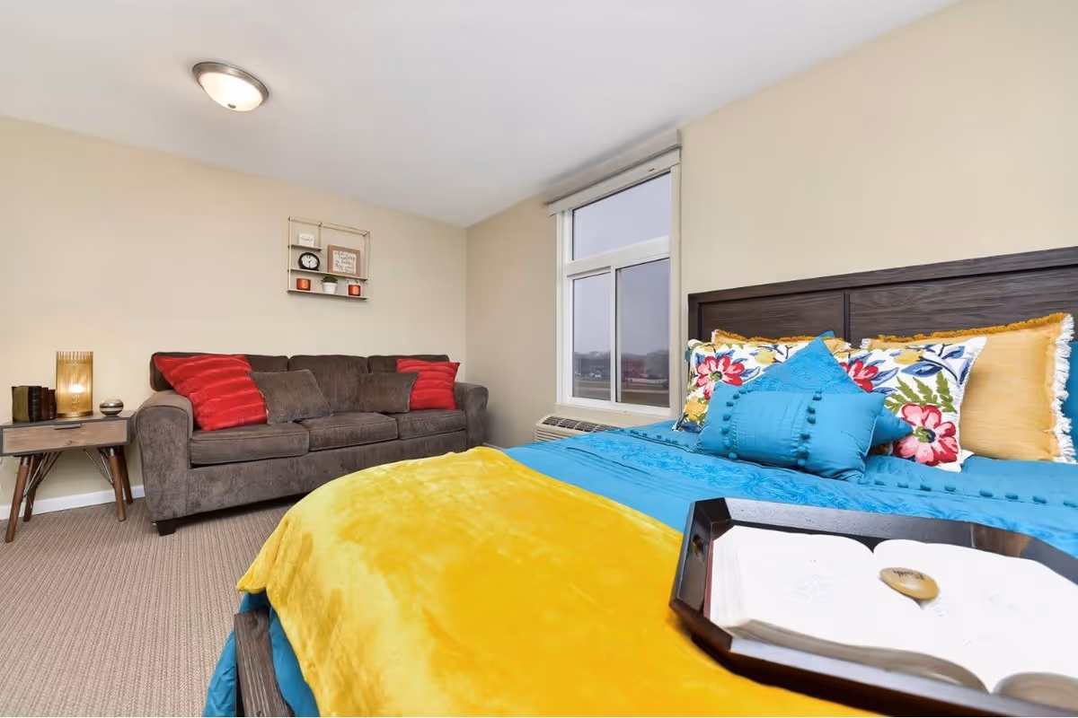 A cozy bedroom in Royal Estates Assistive Living featuring a bed with bright blue and yellow bedding and floral pillows. A tray with an open book and a stone rests on the bed. In the background, there is a brown sofa with red cushions, a small side table with a lamp and books, a window with a view outside, and a small wall shelf with decorative items.