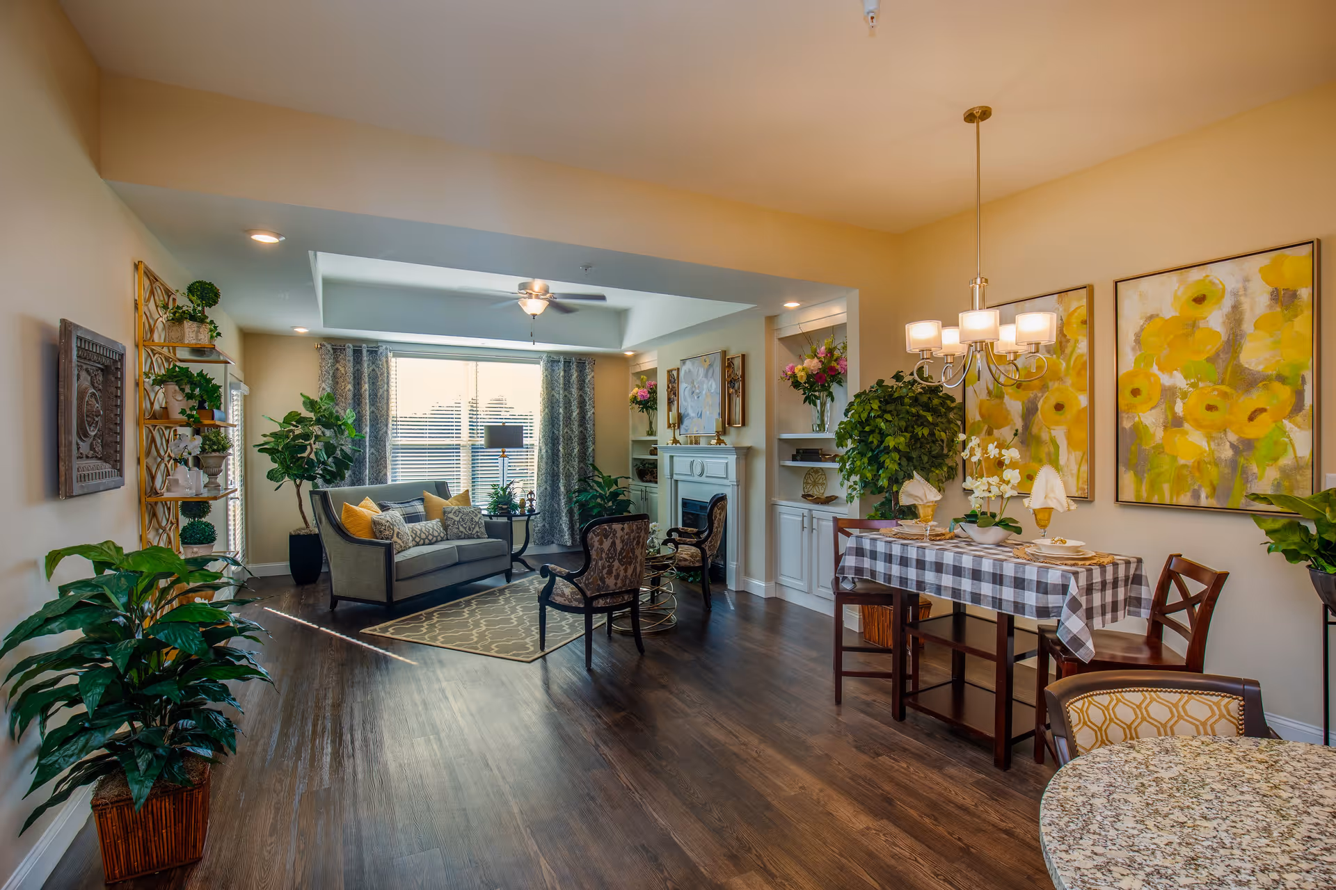 A bright and spacious living and dining area with wooden flooring. The living room features a gray sofa with yellow and patterned cushions, two armchairs, a patterned rug, and a ceiling fan. Large windows with patterned curtains allow natural light to fill the room. The dining area has a table covered with a checkered tablecloth, set with plates and napkins, and is illuminated by a chandelier. The walls are decorated with large floral paintings and there are several green plants placed around the room, creating a warm and inviting atmosphere.