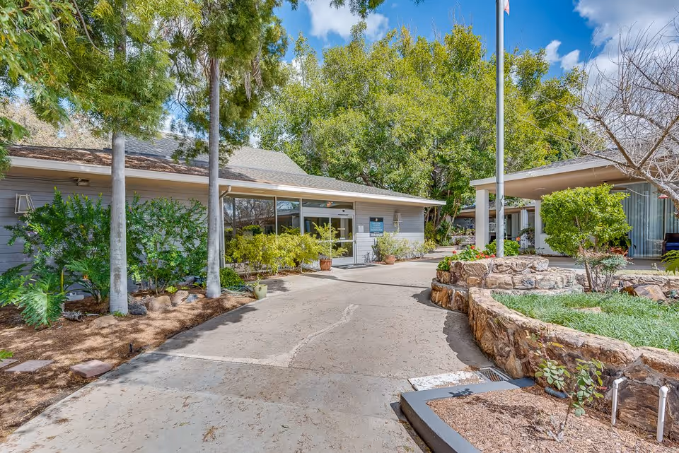 Exterior view of Monte Vista Village facility showing a paved driveway leading to the entrance surrounded by green trees, shrubs, and landscaped garden beds with stone borders under a partly cloudy sky.