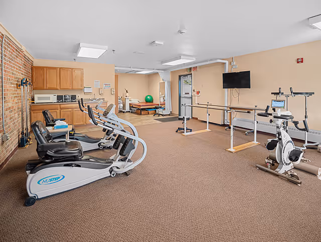 Physical therapy/exercise room with recumbent bikes, parallel bars, and a treatment table.
